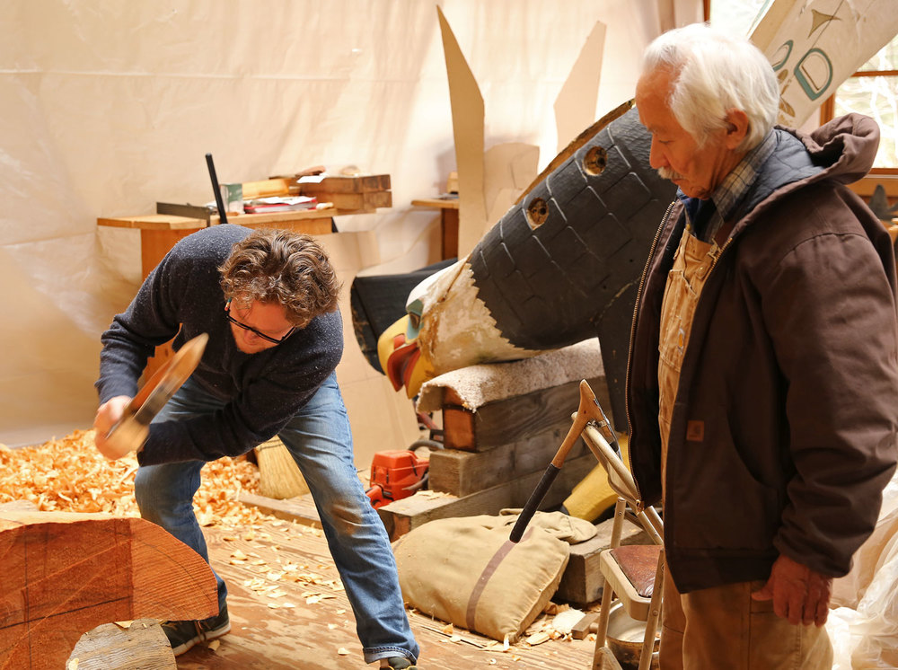 Donald Varnell uses an adze to refine a form as Nathan Jackson watches. They are reproducing the eagle carving "Thundering Wings" in the background. The eagle's wings were removed to transport it to the carving shed.